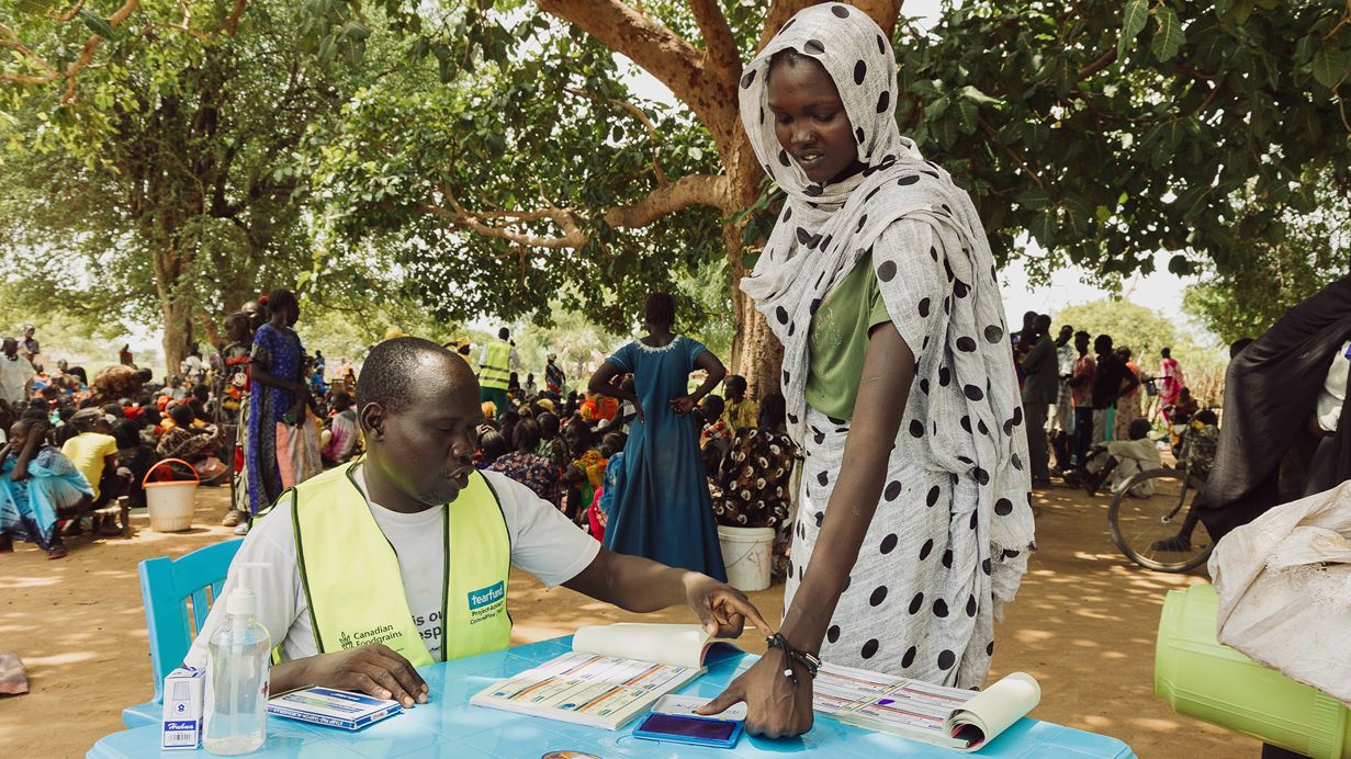 A man in a neon yellow vest sits at a blue table with a woman in a polka-dot headscarf, both looking at and pointing at documents. Many people are gathered in the background under a large tree.