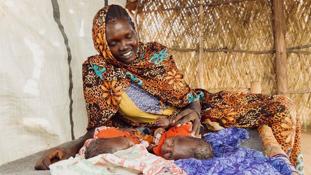 A smiling woman in a colourful, patterned headscarf and dress looks down at two newborns lying on blankets in a simple shelter.