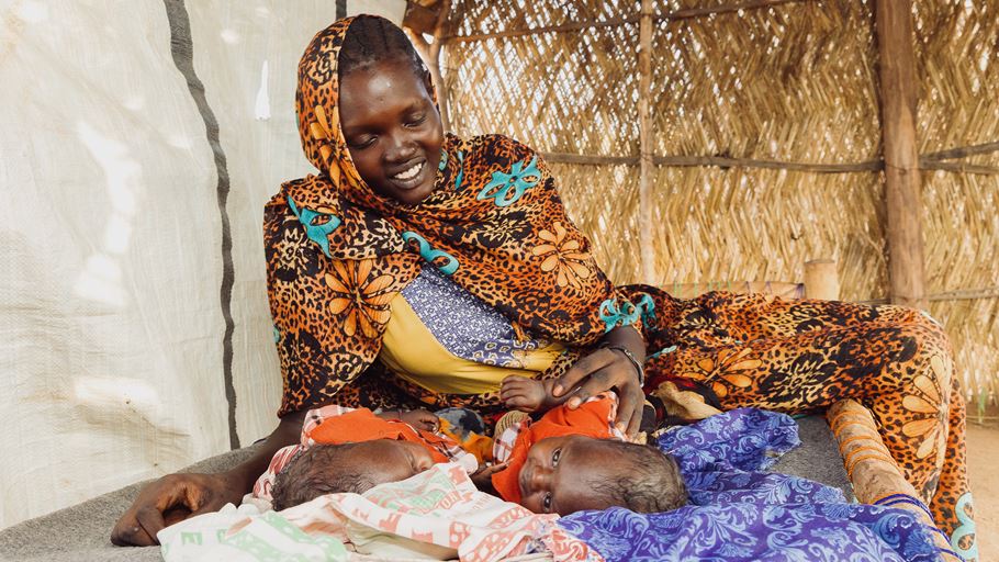 A smiling woman in a colourful, patterned headscarf and dress looks down at two newborns lying on blankets in a simple shelter.