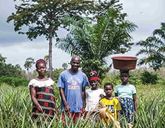 A family stand together in a line, surrounded by green bushes and trees behind them. One of the children balances a large bowl on her head.