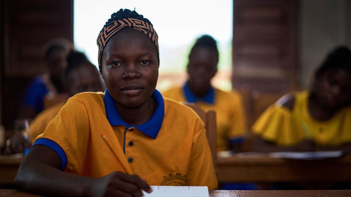 Portrait of a young woman in school uniform, sitting at a desk with a book with other students behind her.