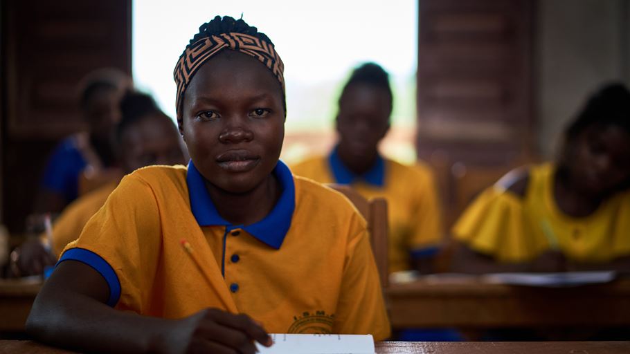 Portrait of a young woman in school uniform, sitting at a desk with a book with other students behind her.