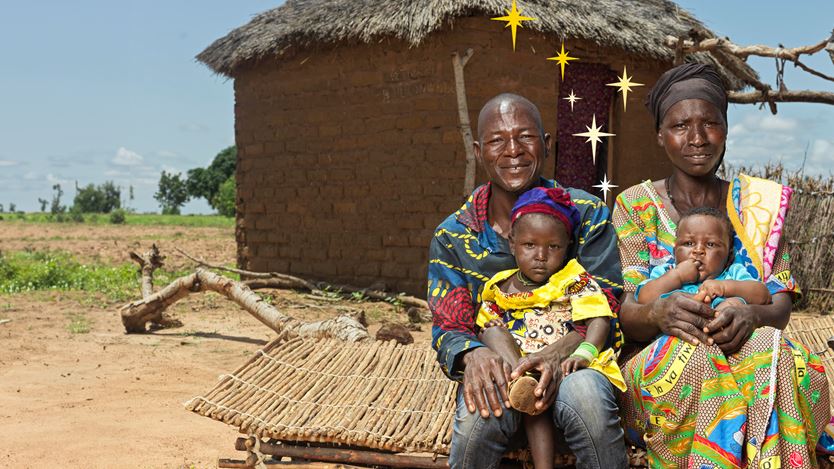 A man and woman are smiling and holding two small children outside a hut.