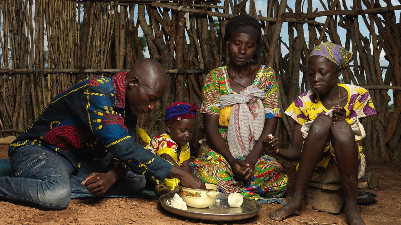 A man, woman, and two children are sitting inside a small hut sharing some bread