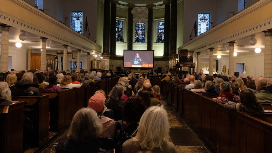 Large crowd in pews watches speaker on screen in church, stained glass and tall columns.