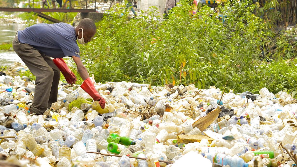 A man wearing red gloves and a face mask picks up plastic waste from a large pile of rubbish near a river, with greenery and a footbridge in the background.