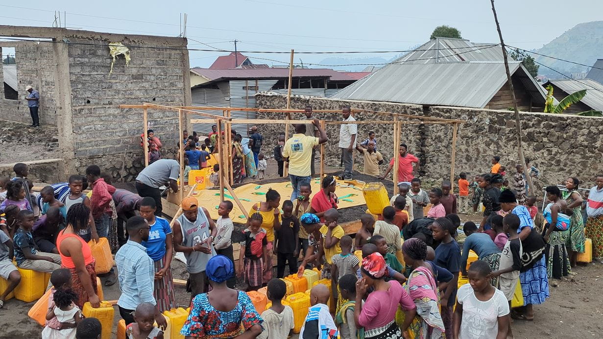 A large group of people including adults and children stand around bright yellow water containers. Some people are building a temporary structure with a wood frame.