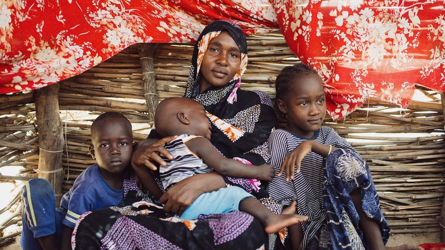 A mother cradles her baby as two other children sit beside her in a rustic shelter.