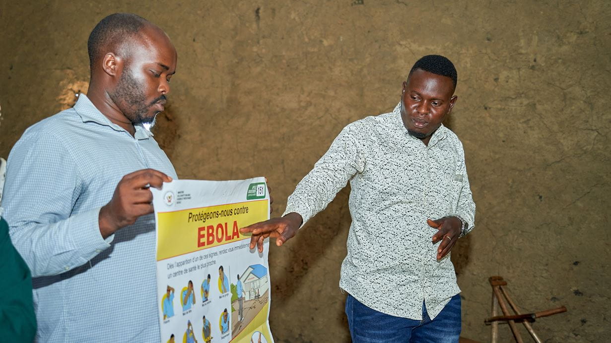 One man holds an Ebola information poster while another gestures to it.