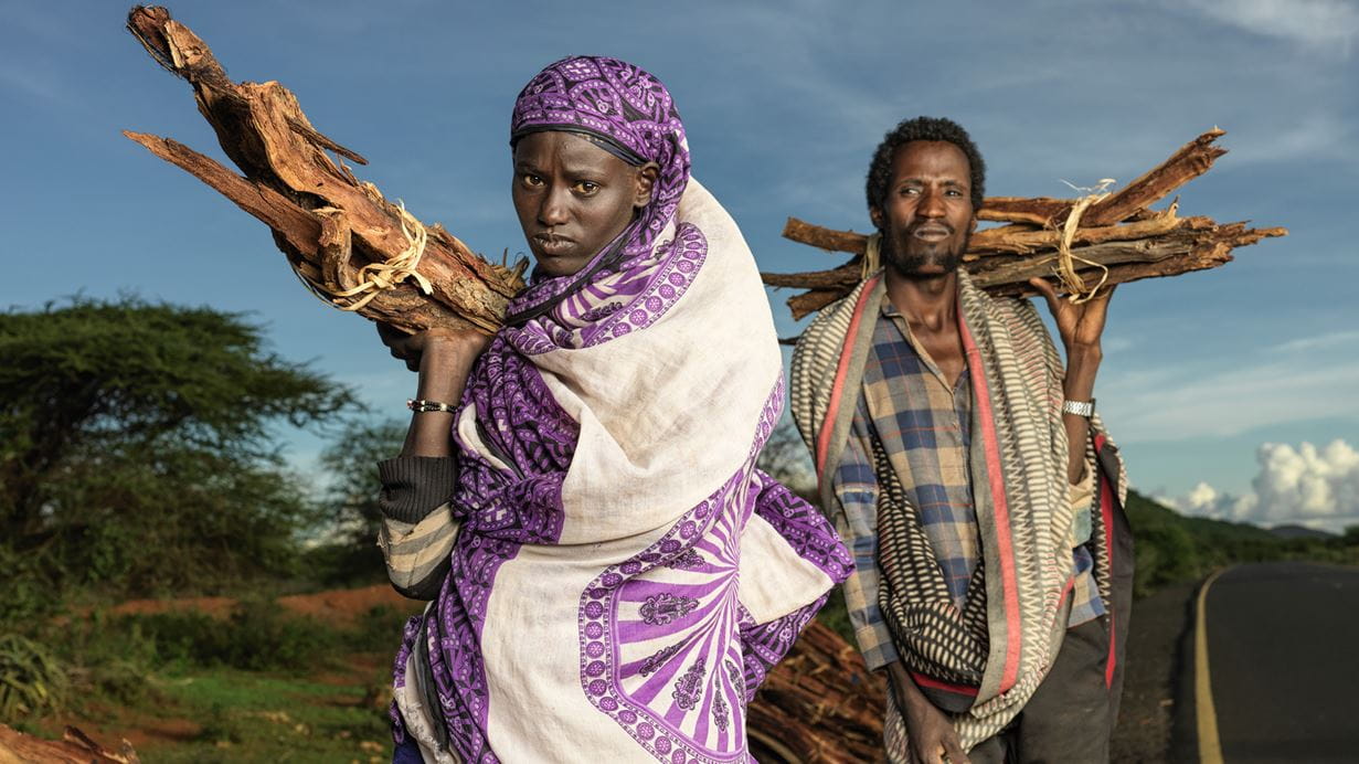 A woman and man walk down a road holding bound piles of wood