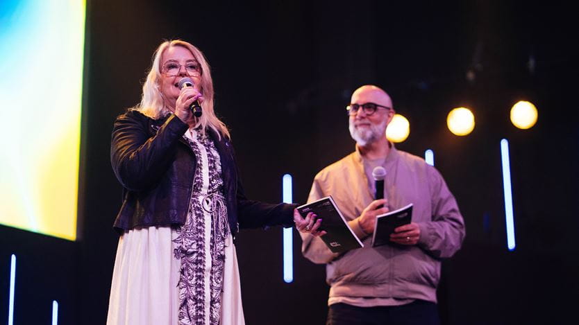 A woman wearing a dress and a man in a jacket stand on a stage under bright lights. The woman speaks into a mic.