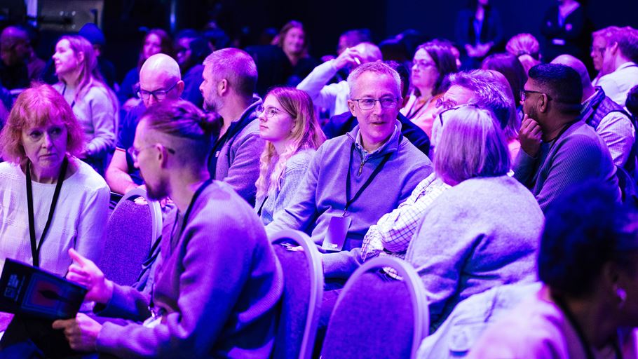 Audience members sit attentively at a conference under bright purple lighting.