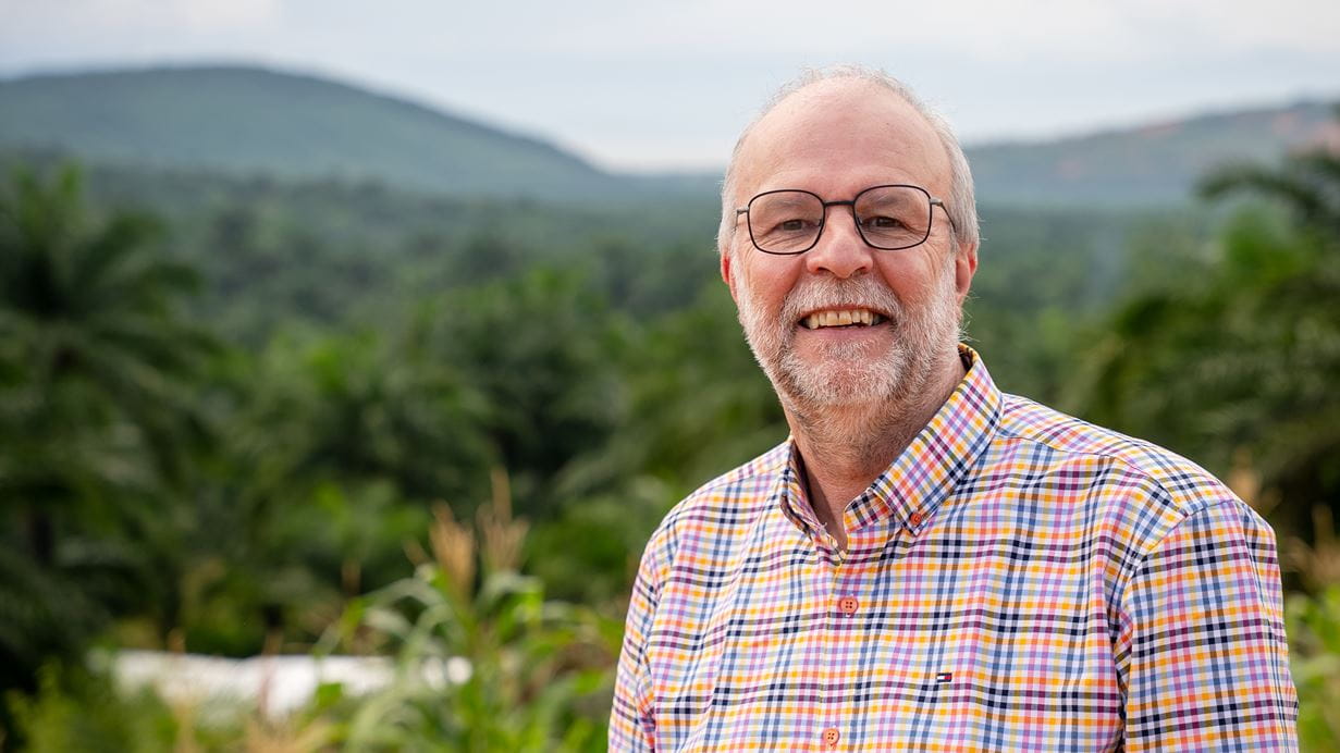 Smiling man with glasses in a plaid shirt, set against a tropical jungle backdrop.