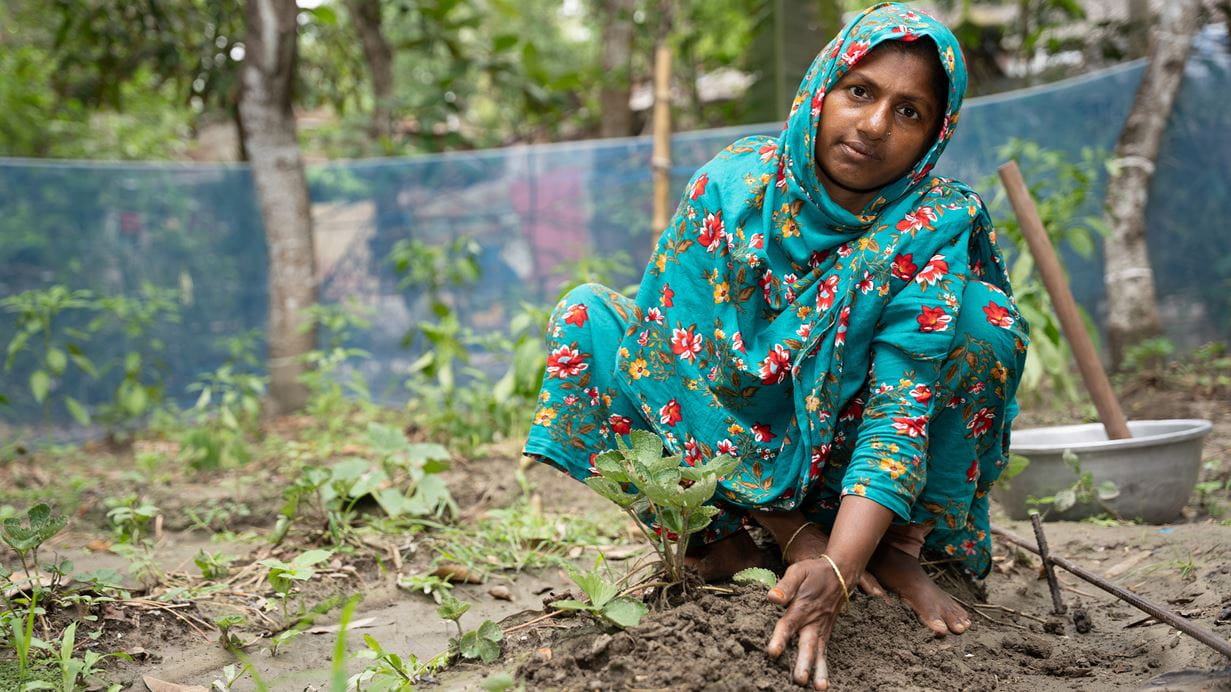 Woman squatting in a garden, tending to a small plant with her hands. She wears a teal floral headscarf and matching dress. Green plants grow in the soil with a blue mesh fence in the background.