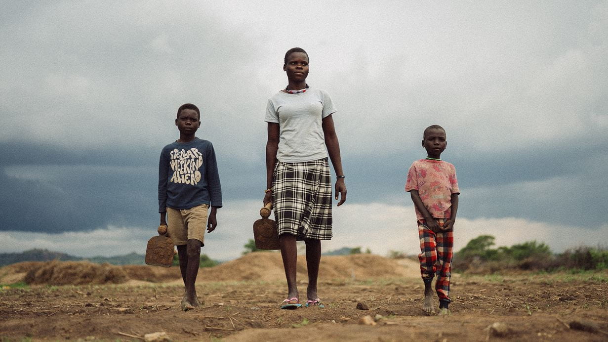 Three children walk along a field with dark cloudy skies behind them. Two of the children hold tools in their hand.