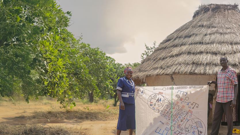 A woman in a blue outfit and a man in a checkered shirt stand outside a traditional thatched hut, holding a large hand-drawn community map. Green trees and dry earth surround them under a cloudy sky.