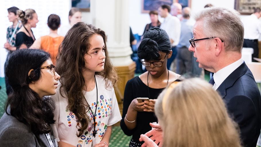 A small group of young women are stood listening to a older man who is talking and gesturing with his hands.