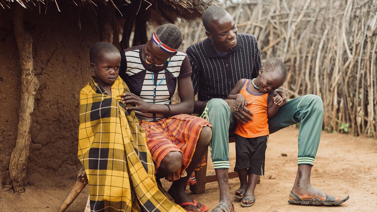A family of four sits outside a traditional dwelling made of mud and sticks. An adult man and woman smile, with a young child in an orange top leaning against the man, also smiling. Another child, wrapped in a yellow and black plaid blanket, looks towards the viewer.