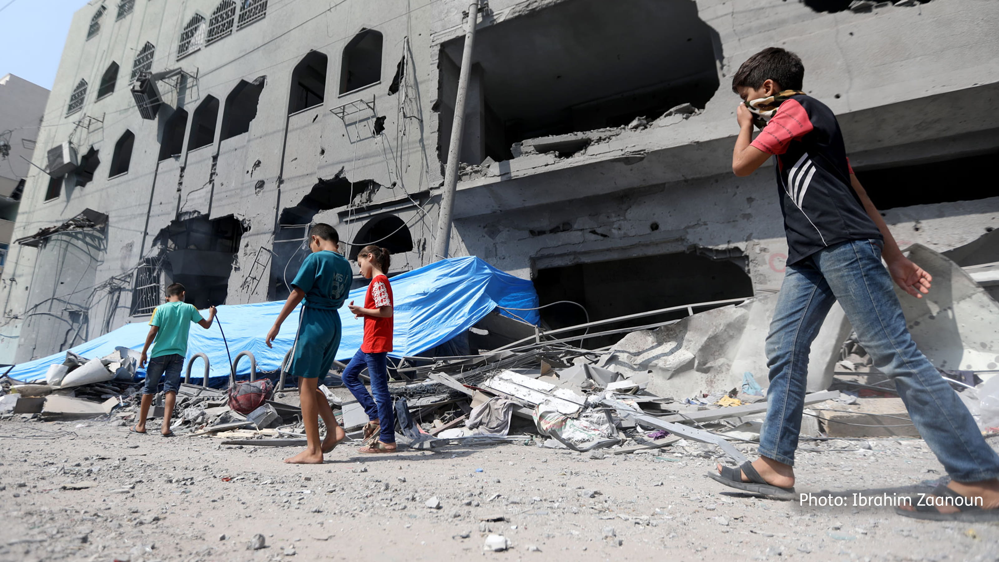 Four children walking past a large damaged grey building with debris on the ground