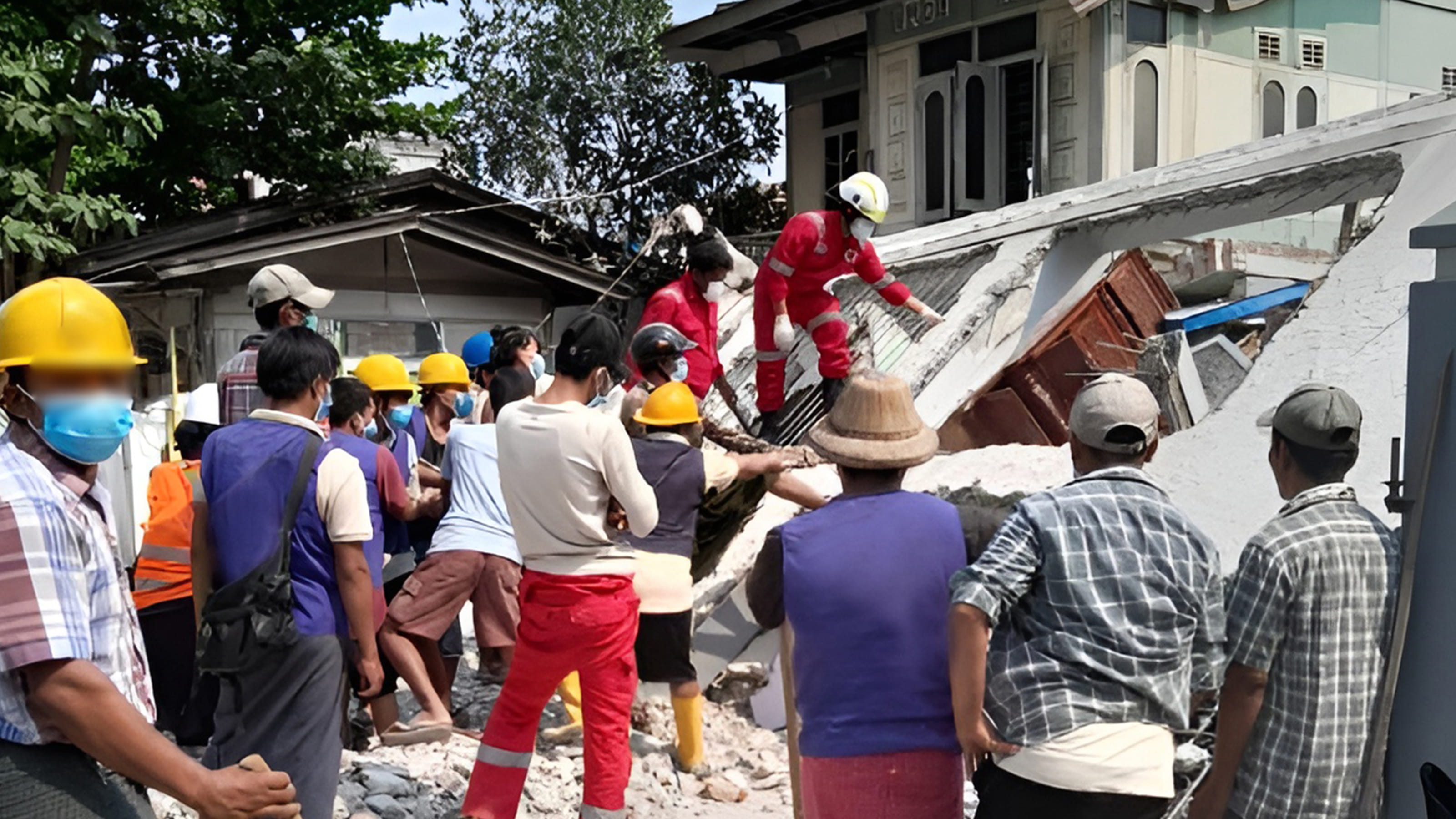 A large group of people gather round a building which has fallen to the ground, some wearing hardhats and masks.