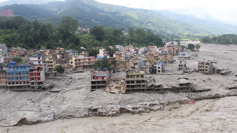 Many multi-storey buildings in Nepal are partially buried in grey mud and debris, with several collapsed structures, alongside a wide muddy river. Green hills and mountains rise in the background.