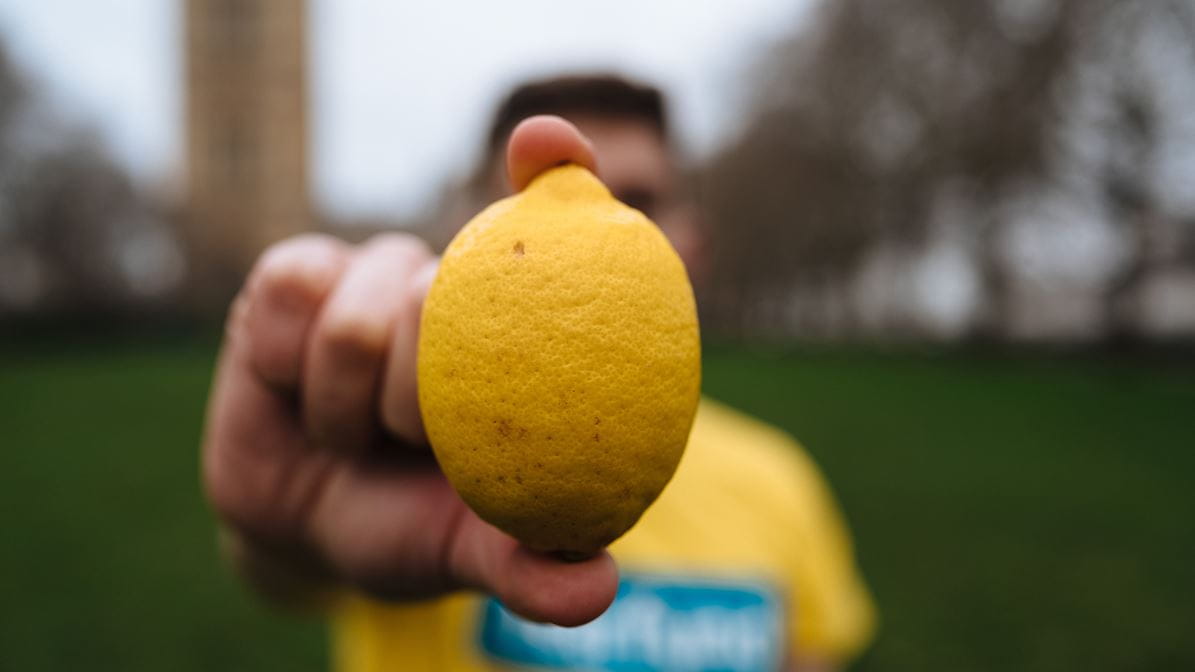 A man in a yellow Tearfund t-shirt stands in front of the Houses of Parliament in London, UK holding a lemon. Image: Tom Price/Tearfund.