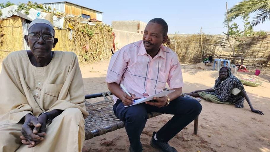 Man interviewing older adults outside in a village setting with simple housing.