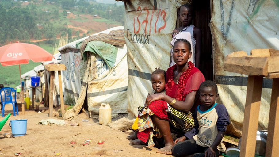 A woman sits in front of a temporary shelter with a young child sat on her lap. Next to her are two other children. 
