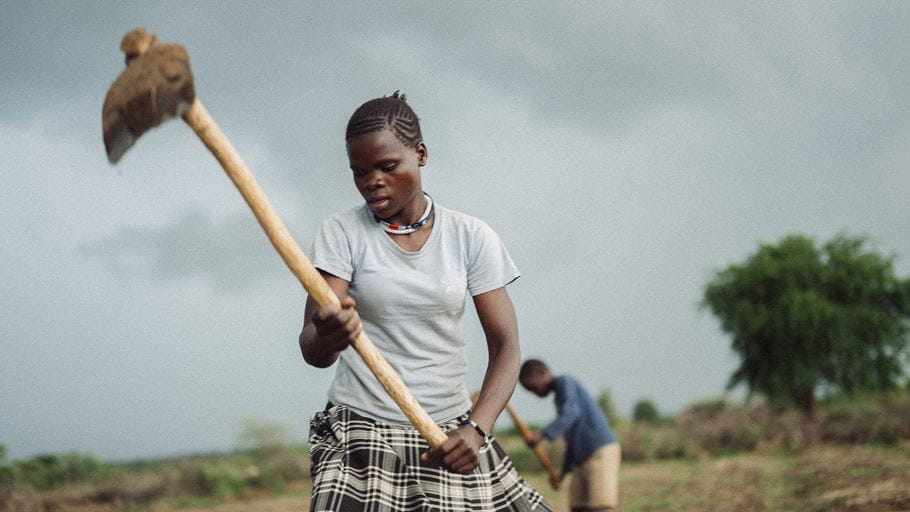 Woman with a hoe tilling a field under a cloudy sky; another person works in the background.