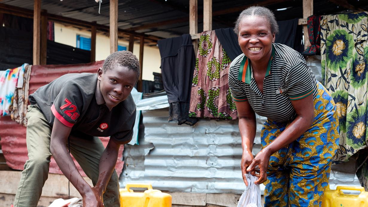Smiling woman and boy washing in a rural outdoor setting with laundry hanging behind.