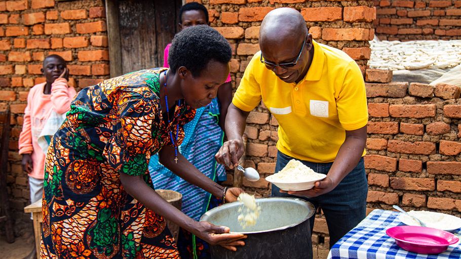 A woman in a colorful patterned dress and a man in a yellow shirt serve food from a large pot into plates, standing in front of a red brick wall. Others observe in the background.
