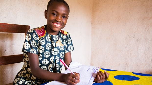 A smiling girl sits at a table with a colorful cloth, holding a pen and writing in a notebook, wearing a patterned dress in a simple indoor setting.