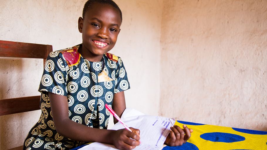 A smiling girl sits at a table with a colorful cloth, holding a pen and writing in a notebook, wearing a patterned dress in a simple indoor setting.