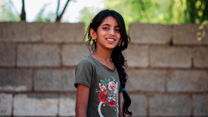 A smiling girl with a long braid stands outdoors in front of a stone wall, wearing a green shirt with floral patterns, with trees visible in the background.