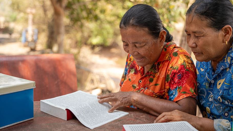 Two women are sat at a table outside reading from a bible