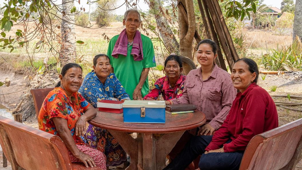 A group of six women are sat and stood around a wooden table outside. On the table are bibles and a metal tin. 