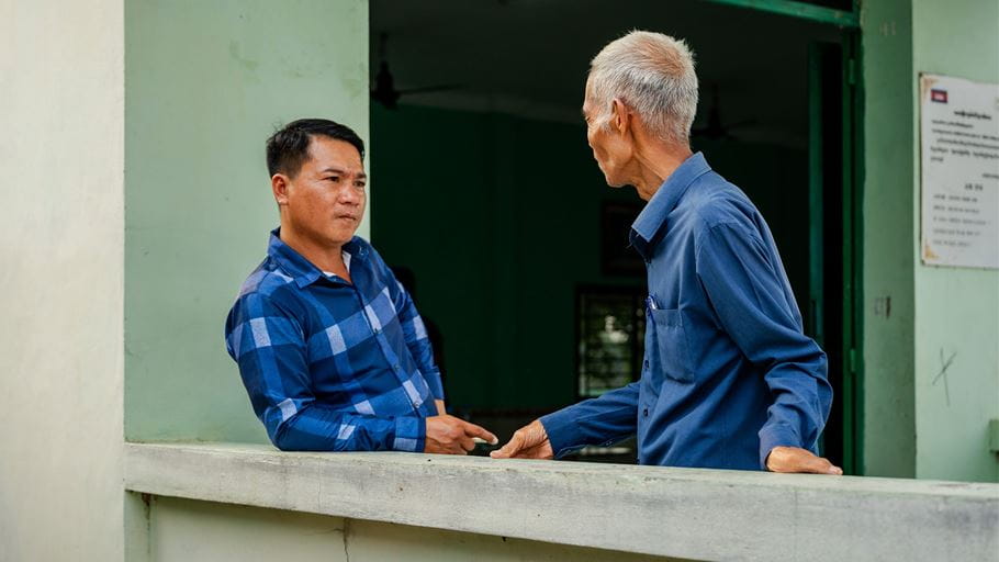 Two men are stood outside a doorway. One man leans against a wall and the other is looking at him, with his hands on the wall.