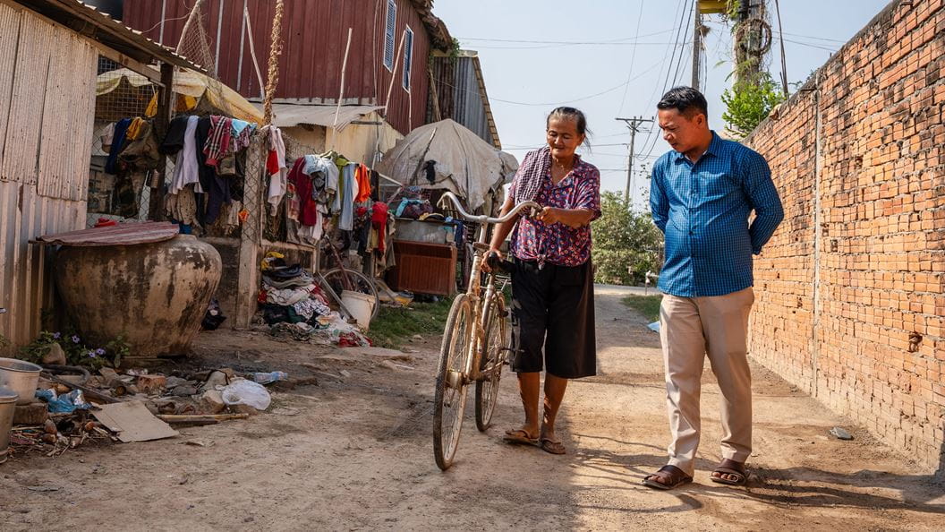 A woman pushing a bicycle and a man are walking down a narrow street, between a wall and buildings.