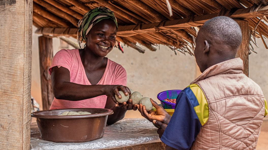 A woman stood in a small wooden market stall smiles and passes several soaps from a bowl to a man in front of her.