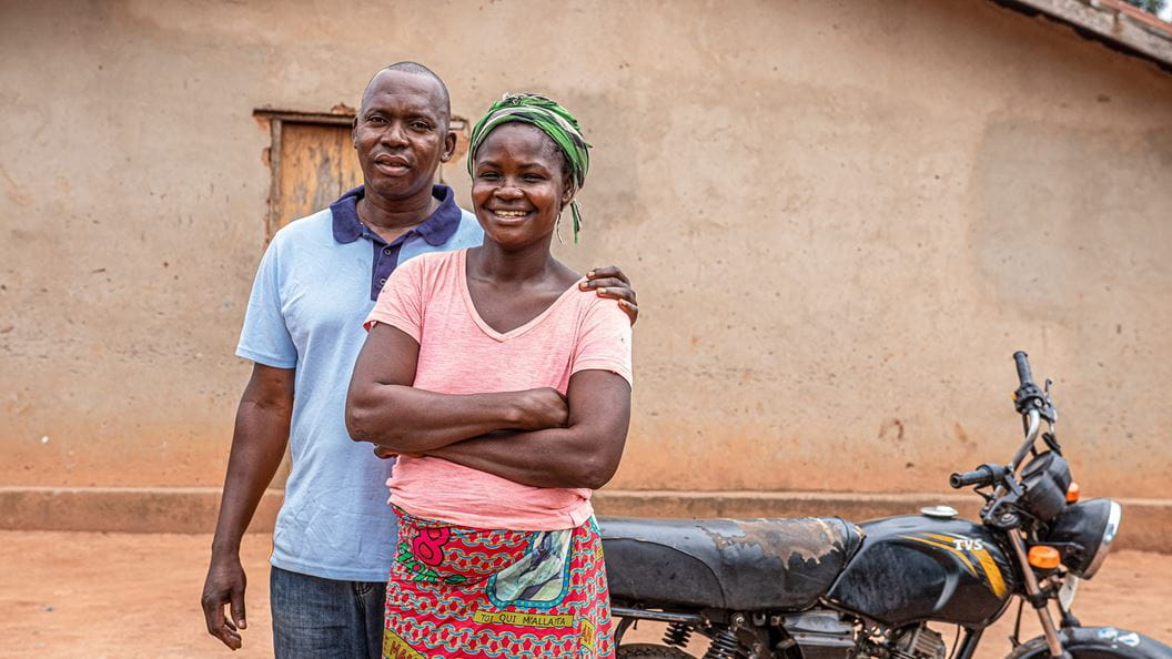 A man and woman stand in front of a small motorbike. The man has his arm around the woman.