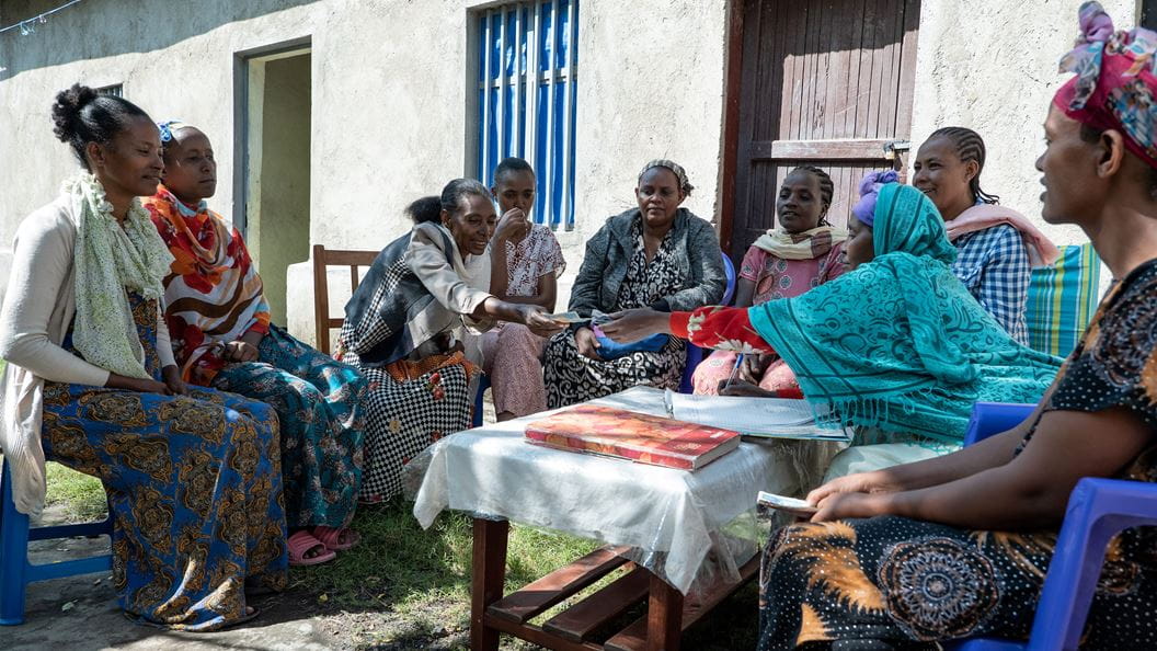 A group of women sitting outside around a table with a building in the background