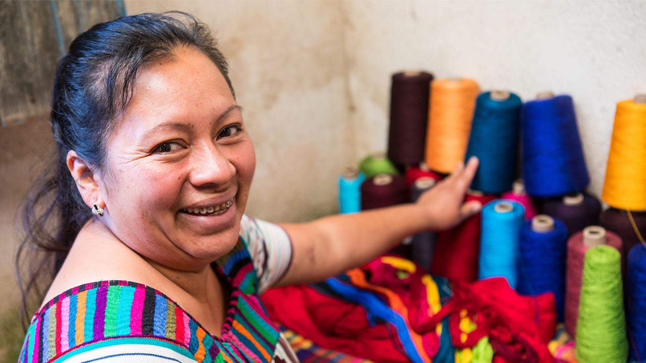A woman in a colourful dress, smiling and pointing at a selection of different coloured spools of thread 