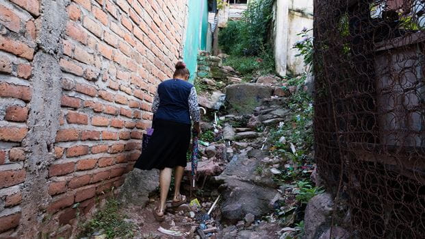 A woman walks up a rocky path alongside a brick wall with an umbrella in hand.