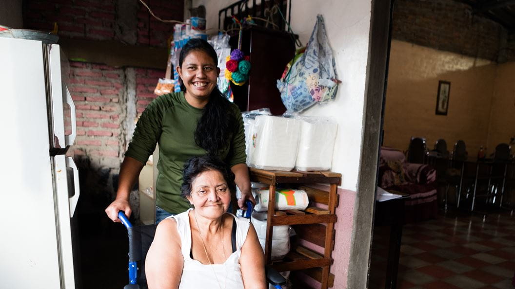 Two women, one in a wheelchair, smile in a home setting with brick and personal items around.