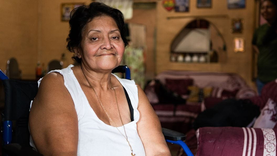 Portrait of a senior woman sitting in a wheelchair indoors, looking at the camera and smiling slightly.