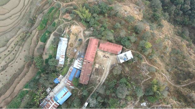 Aerial view of buildings below, terraced fields, and dirt paths on a green and brown hillside.