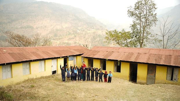 Children and a teacher raise hands in front of school buildings in a rural mountain setting.