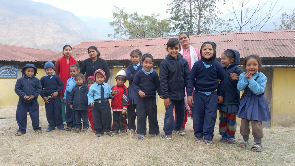 Group of smiling children and adults standing outside a school with mountains in the background.
