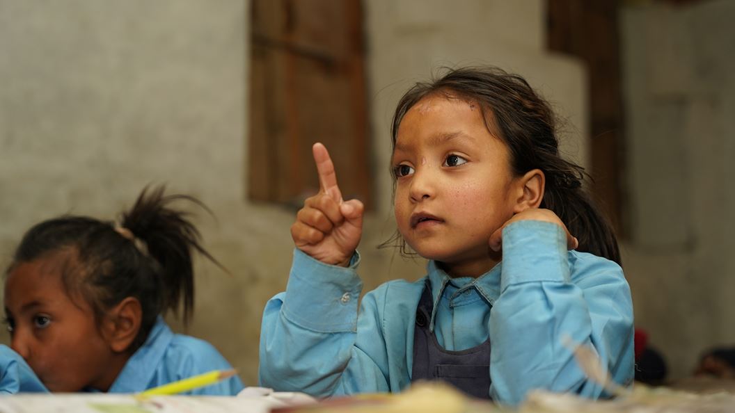 Young girl in blue school uniform raises hand in classroom, another student behind her.