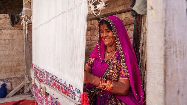 A Pakistani woman dressed in a bright pink patterned dress and head scarf smiles as she works at a weaving loom.
